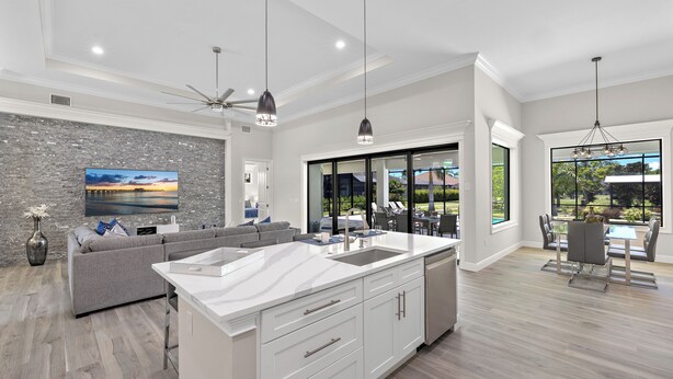 A modern kitchen with a large island, a dining area with a table and chairs, a gray sofa, and a view of the outdoors through large windows.