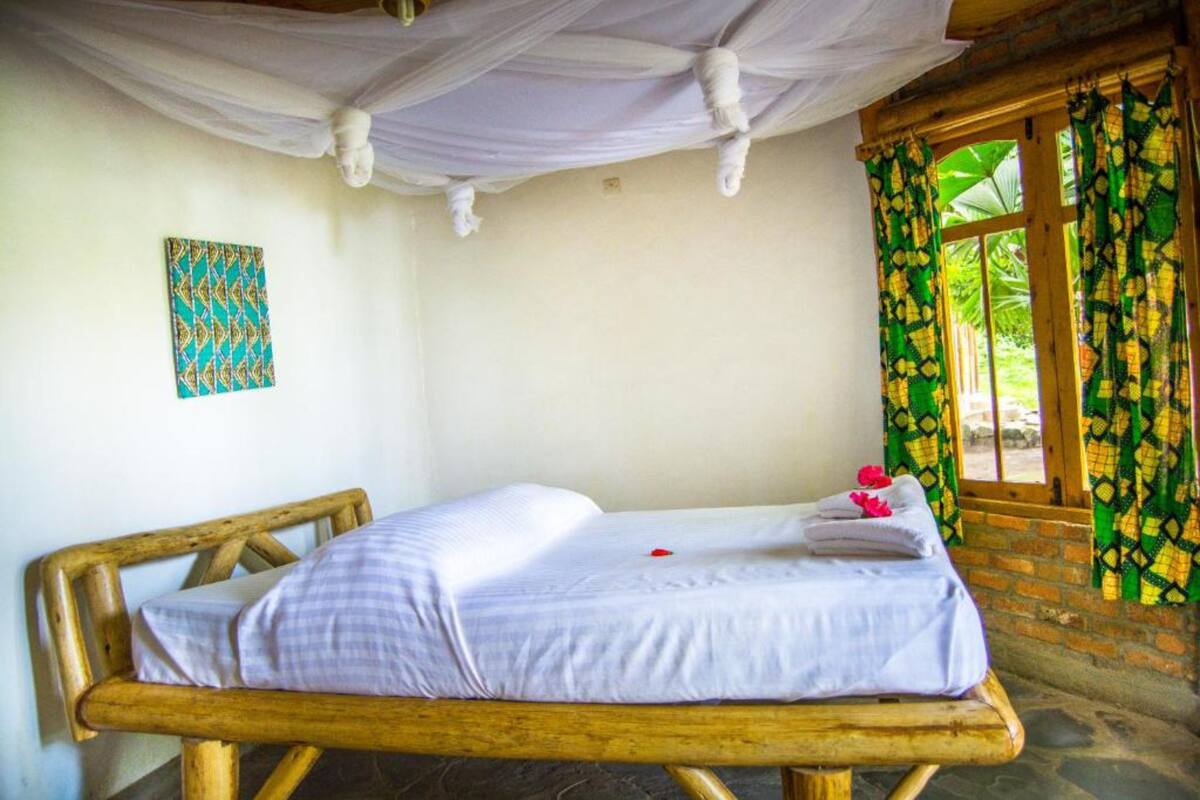 A single bed with a white mattress and a wooden frame, a wall with a geometric patterned artwork, and a window with green and yellow curtains.