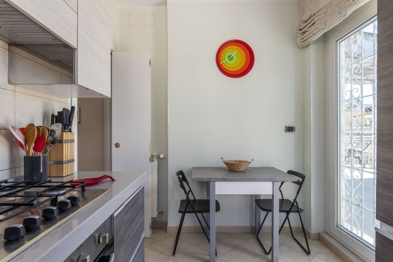 A kitchen with a gas stove, a dining table with two chairs, a wall clock, and a window with a metal grill.