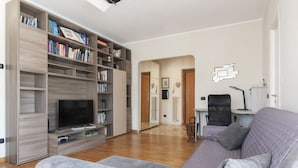 A living room with a large bookshelf, a flat-screen TV, a grey sofa, and a dining area with a table and chairs. - GuestHost - Cozy and comfortable 75sqm apartment, able to accommodate up to 6 people, located on the 5th floor with elevator (not suitable for disabled peolple) of a building less than 10 minutes walk away from Bologna Metro stop and the homonymous squ (Roma)