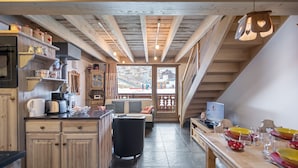 A cozy kitchen with wooden beams, a dining area with a table set for four, and a view of a snowy landscape through a large window.