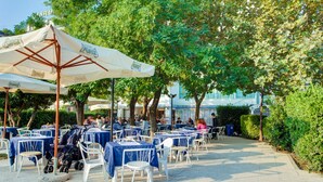 An outdoor cafe with tables and chairs, covered by umbrellas, and surrounded by trees. - Hotel Eritrea (Cesenatico)