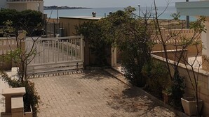 En stenlagd innergård med en grind, planterade blommor och utsikt över havet och avlägsna byggnader.