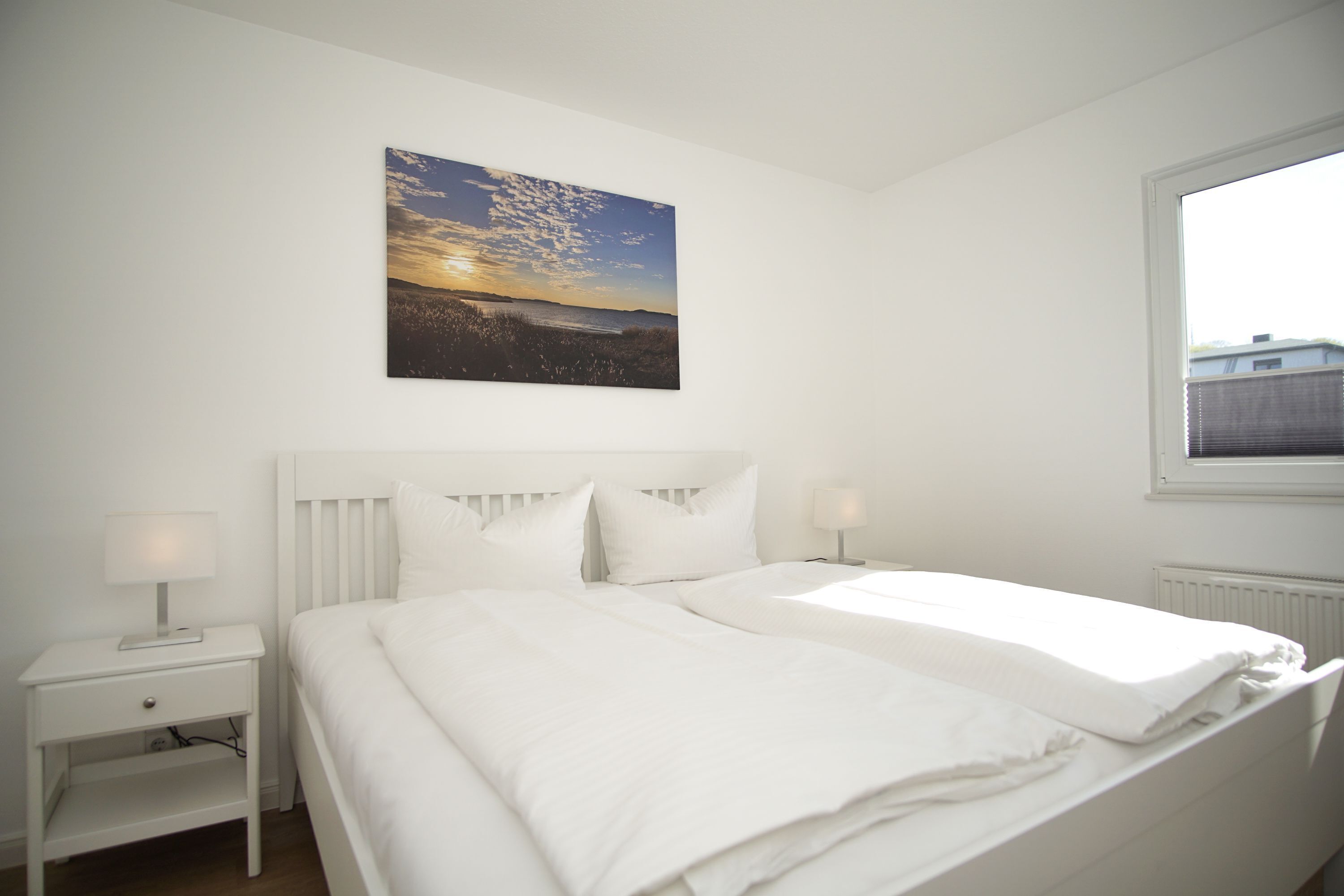 Ein Schlafzimmer mit einem Bett, einem Nachttisch, einer Lampe, einem Fenster und einem Wandbild.