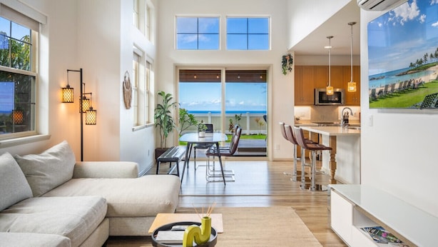 Ein geräumiges Wohnzimmer mit großem Fenster und Balkonblick, eine Küchenzeile mit Barhocker und ein Essbereich mit Meerblick.