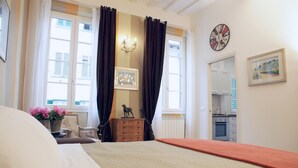 Et soveværelse med en seng, et kommode, et lille bord med blomster, en lampe, et ur og udsigt til køkkenet gennem en åben dør.