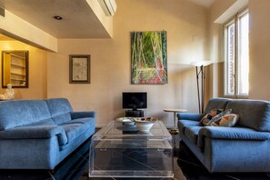 Een woonkamer met twee blauwe banken, een glazen salontafel, een schilderij, een televisie en een vloerlamp.