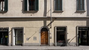 Eine Gebäudefassade mit einer zentralen Holztür, Schaufenstern mit den Aufschriften "EBY'S" und "SAN PIERINO" sowie einem heruntergelassenen Rollo auf der linken Seite.