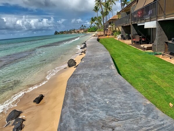 A beachfront property with a concrete walkway leading to a grassy area and a pool, overlooking the ocean with palm trees and a building with outdoor seating.