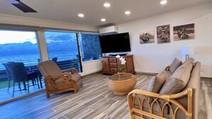 Amplia sala de estar con un gran ventanal con vista al mar, un ventilador de techo, un televisor de pantalla plana y un sofá de mimbre.