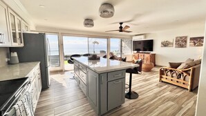 Amplia sala de estar con cocina, comedor y un televisor grande. Cuenta con ventilador de techo, vista al océano y balcón.