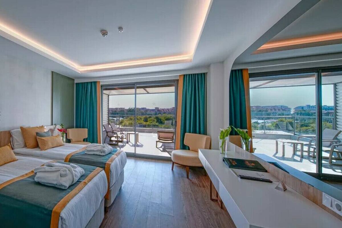 A modern hotel room with two beds, a desk, a balcony, and a large window offering a city view.