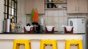 Een keuken met een fornuis, een spoelbak en gele krukken. Boven het aanrecht zijn kastjes en er is een raam met jaloezieën.