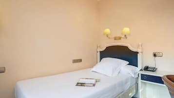 A single bed with a white mattress and pillows, a bedside table with a lamp, and a small drawer.