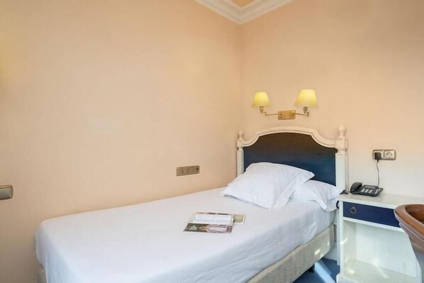 A single bed with a white mattress and pillows, a bedside table with a lamp, and a small drawer. - Hospedium Hotel Sky Blu Sondika (Sondika)