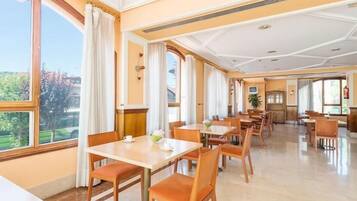 A hotel dining area with multiple tables and chairs, large windows with curtains, and a view of the outdoors.