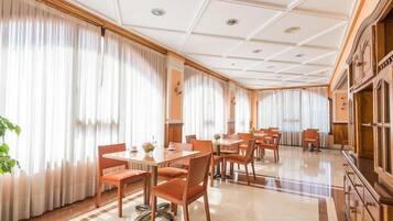 Un comedor amplio con varias mesas y sillas, ventanas grandes con cortinas y un piso pulido.