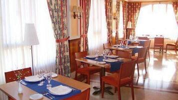 A dining area with tables set for four, each with a blue table runner, white plates, and wine glasses. There are curtains with floral patterns and wall sconces with lamps.