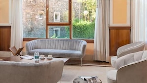 Una sala de estar moderna con un sofá gris, una mesa de centro redonda y un reposapiés gris. Se ve una ventana con vistas a un edificio de piedra y vegetación.