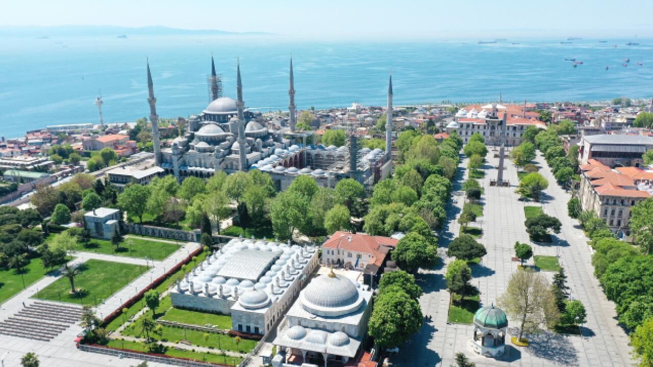 An aerial view of a historic city with prominent domed buildings and minarets, surrounded by greenery and a body of water.