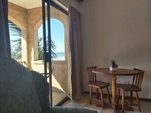 A room with a balcony overlooking a beach, a round wooden table with chairs, and a potted plant on the table.