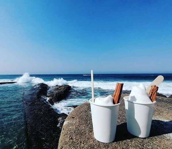 Two white cups with straws and a wooden spoon on a rocky ledge overlooking the ocean.