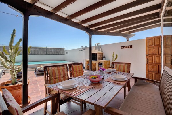 Une terrasse extérieure avec une table à manger pour quatre personnes, entourée de chaises, d’un cactus et, au fond, d’une piscine.