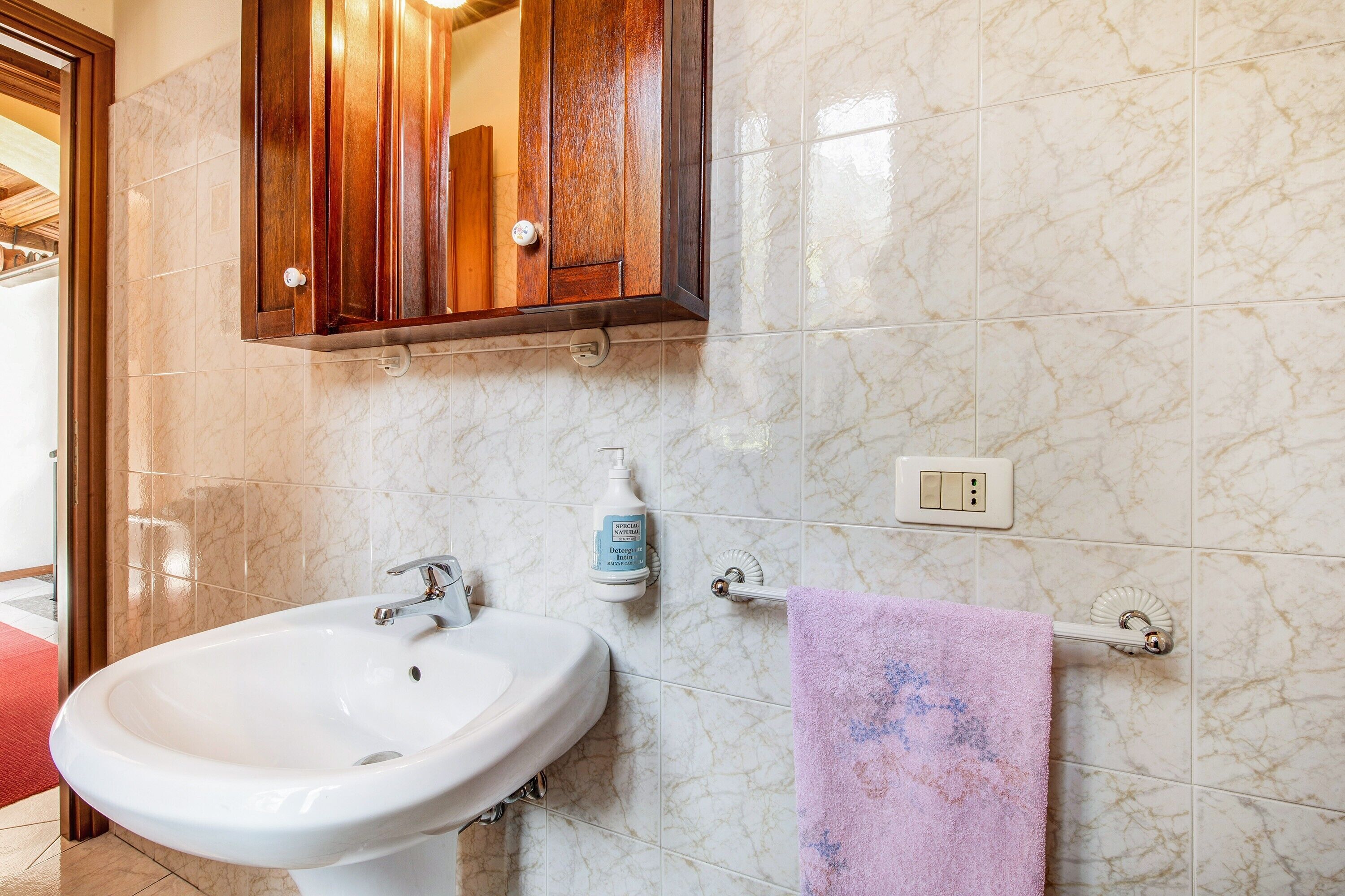Een badkamer met een wit wastafel, een spiegel erboven, een houten kastje, een marmeren wand, een handdoekhouder en een fles handzeep.