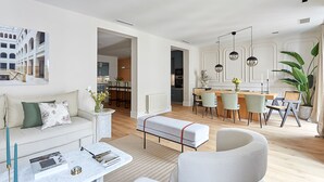 Ett modernt vardagsrum med en soffa, ett soffbord och en pall. Det finns en matplats med ett bord och stolar, och ett kök syns i bakgrunden.