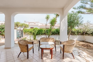 A covered patio area with wicker furniture, a tiled floor, and a view of a building with a tiled roof and greenery.