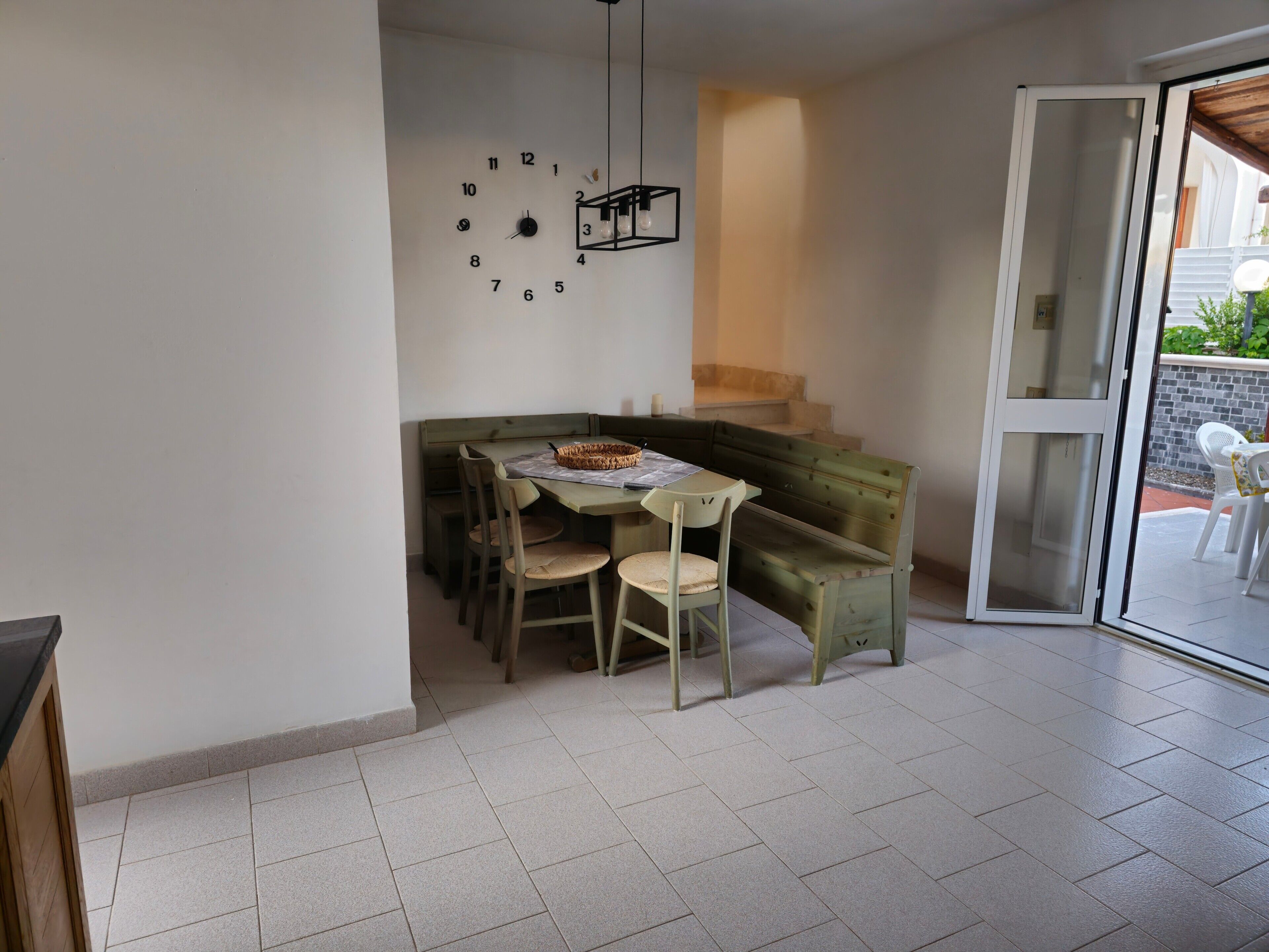 Zona pranzo con tavolo, sedie, una panca, un orologio a parete e una porta scorrevole in vetro che dà sull'esterno.