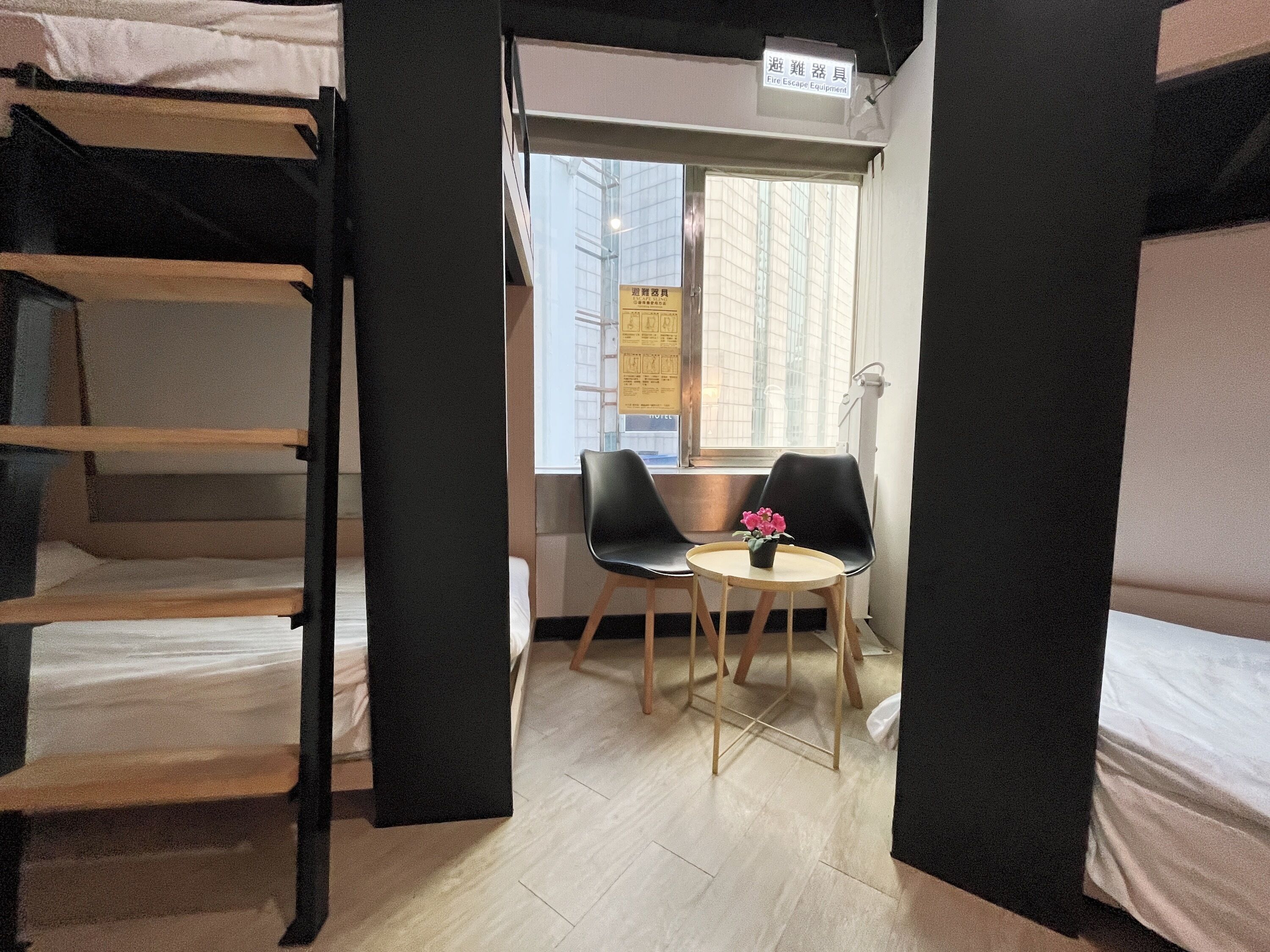 A dormitory room with bunk beds, a small table with a flower, and a window with a view of buildings.