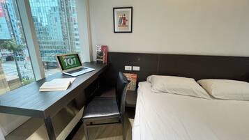 Habitación de hotel con cama, escritorio con computadora portátil, silla, ventana con vista a edificios y lámpara de pared.