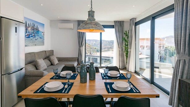 A modern dining area with a wooden table set for two, a sofa, and a large window offering a view of the landscape. - Spacious apartment in Cómpeta with shared pool and jacuzzi (Cómpeta)