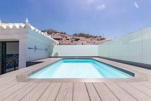 A rooftop swimming pool with a wooden deck, a white wall with a door, and a view of buildings in the background.