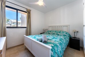 A bedroom with a bed, a nightstand, a window with curtains, a ceiling fan, and a view of buildings outside.