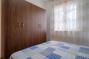 Une chambre avec une grande armoire, un lit recouvert d’une couette à carreaux et une fenêtre ornée de rideaux légers.