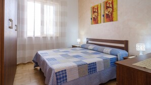 Un dormitorio con una cama, una mesita de noche, una lámpara, un ventanal con cortinas translúcidas y dos cuadros colgados en la pared.
