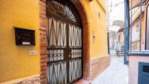 Una calle estrecha con un edificio amarillo, una puerta de metal con diseños decorativos y un buzón con la etiqueta 'POSTA'.