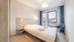 Habitación de hotel moderna con una cama grande, mesitas de noche, una silla y vistas a la ciudad desde la ventana.
