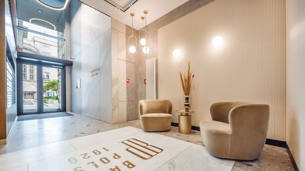 Un vestíbulo moderno con piso de mármol, dos sillones color beige, una mesita auxiliar y un jarrón alto con plantas secas.
