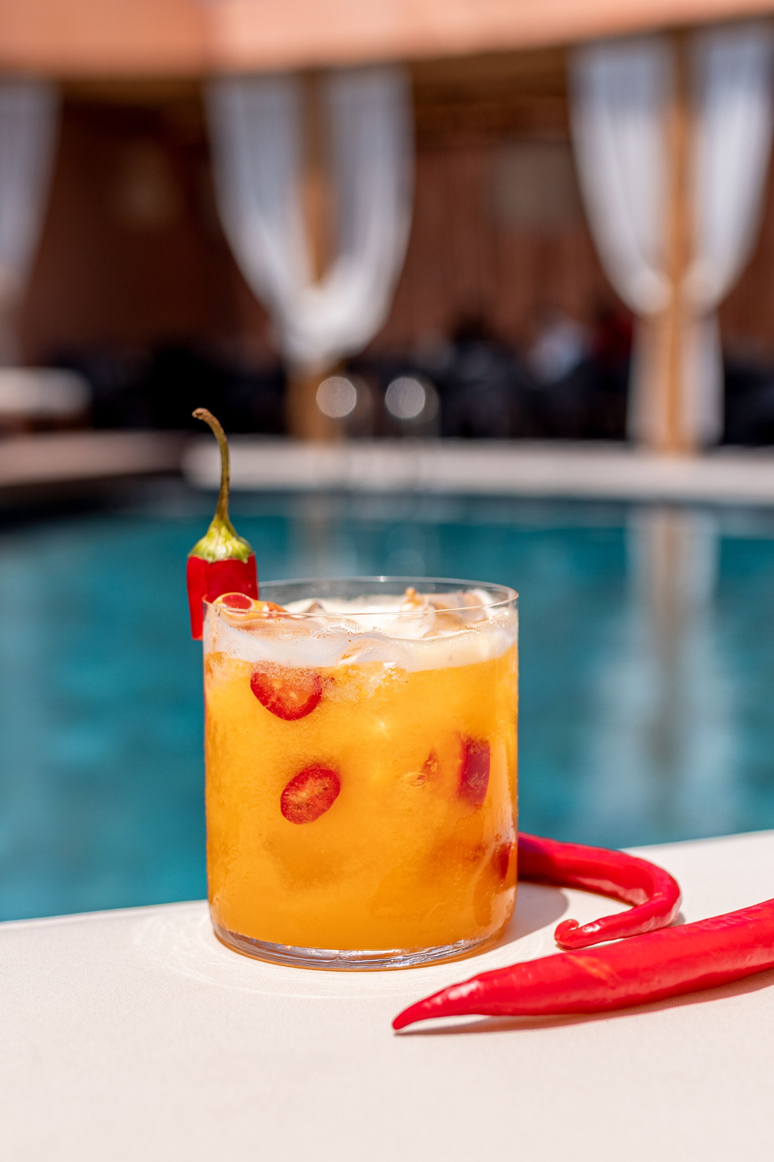 A glass of iced drink with a red chili garnish, placed on a white surface near a pool.