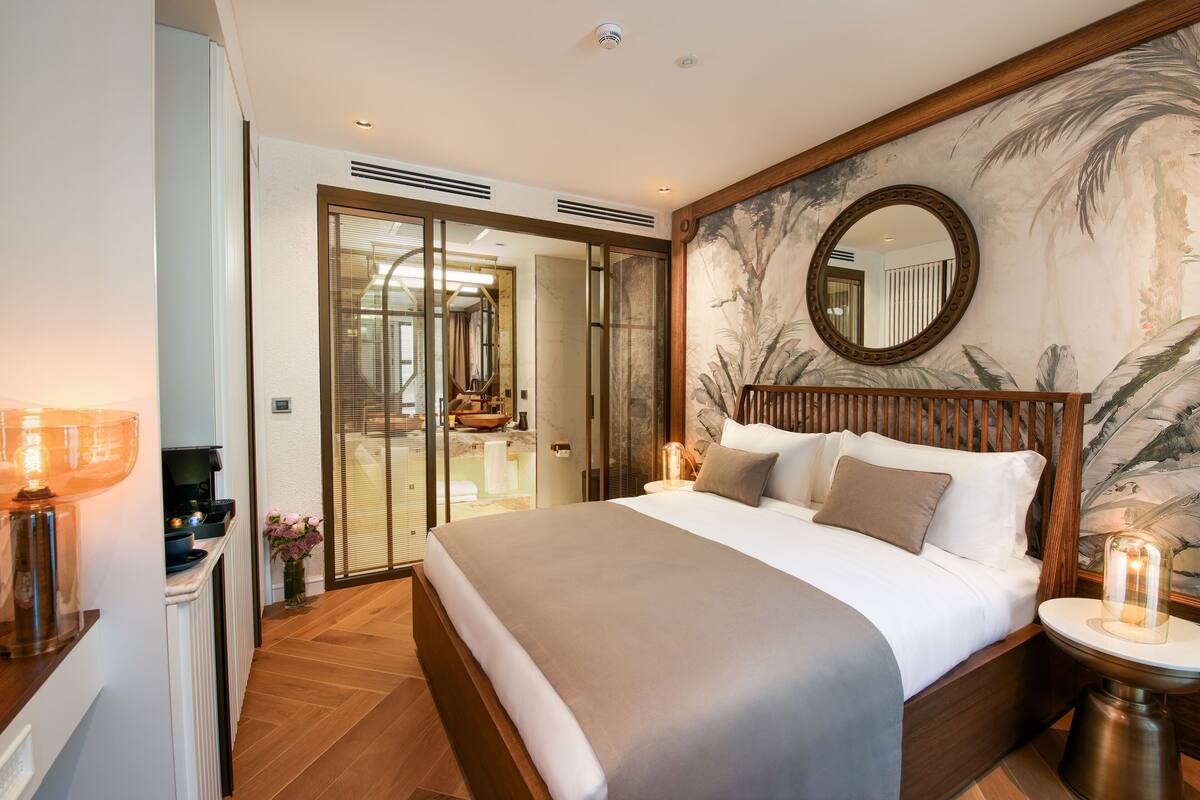 Une chambre d’hôtel moderne avec un grand lit, un miroir, une table de chevet et une vue sur la salle de bain grâce à une porte en verre.