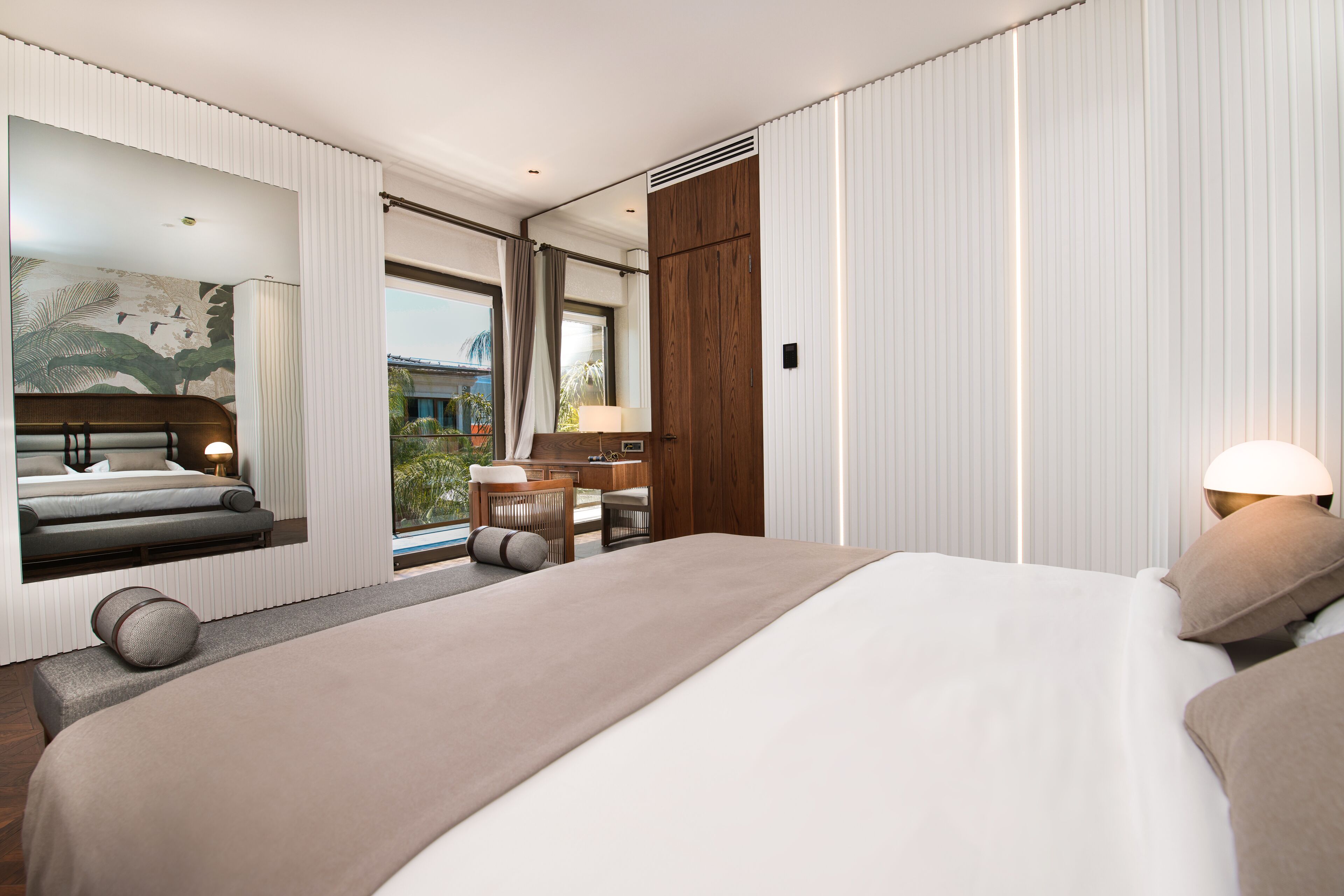 A modern hotel room with a large bed, a seating area with a sofa and chairs, a wooden desk, and a large window offering a view of the outdoors.