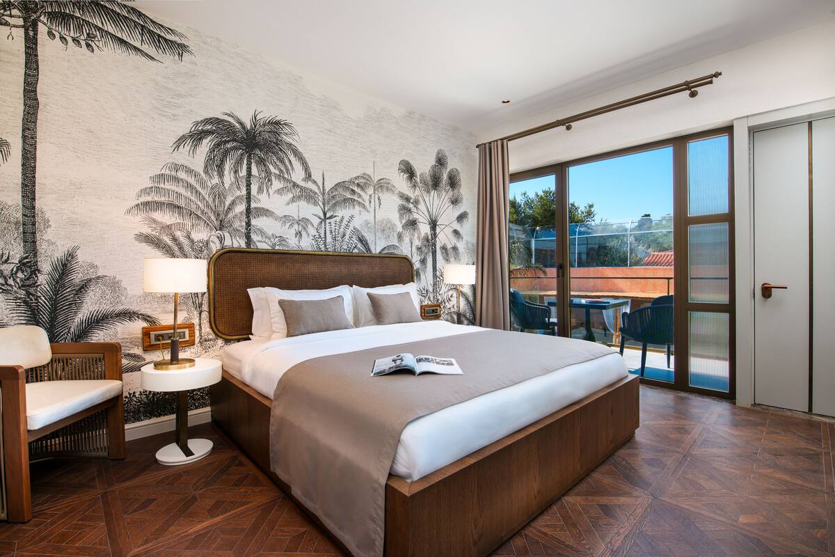 Un grand lit avec une tête de lit en bois, une table de chevet avec une lampe, une chaise et une fresque tropicale sur le mur.