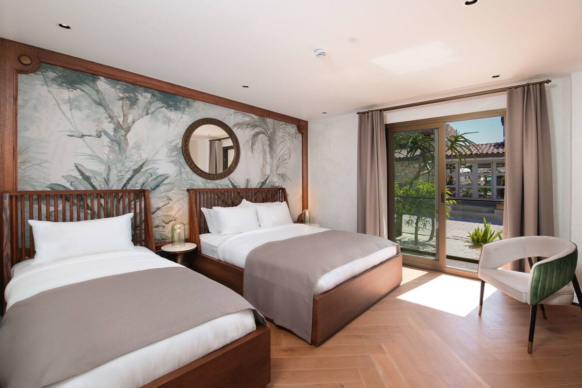 Une chambre d’hôtel avec deux lits, une grande fenêtre donnant sur un beau paysage, une chaise et un mur orné d’un grand miroir rond.
