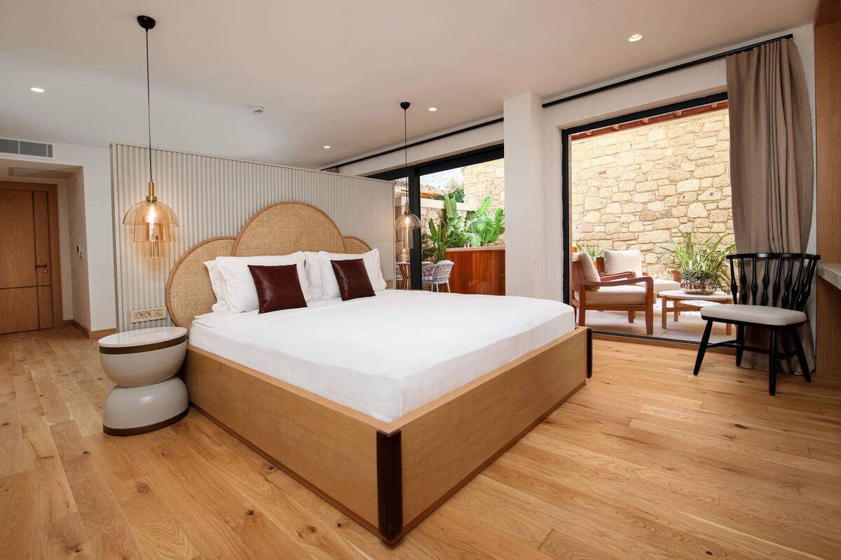Une chambre moderne avec un grand lit, des tables de chevet, un luminaire suspendu, une vue sur l’extérieur et un coin salon avec une chaise et une table.