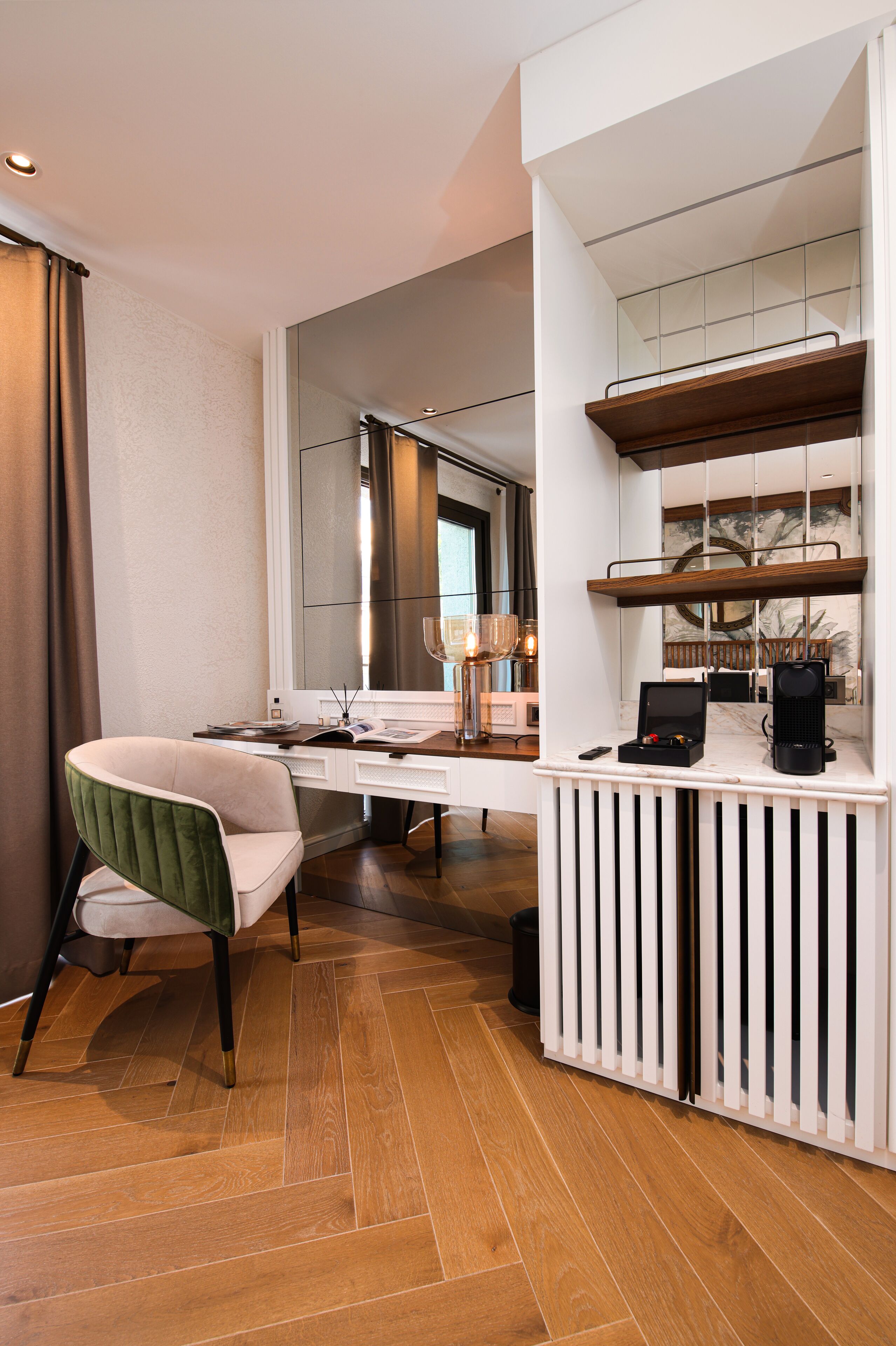 A modern hotel room with a wooden floor, a desk with a chair, a large mirror, and a coffee machine.