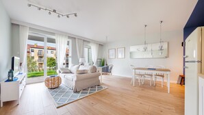 A modern living room with a sofa, dining table, and chairs. There is a large window with curtains, a hanging light fixture, and a mirror on the wall. - Apartamenty Sun & Snow Baltic Sun (Sztutowo)