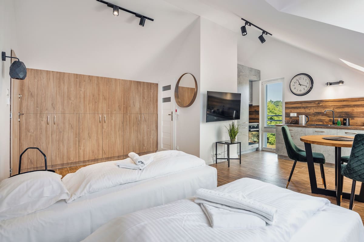Ein modernes Schlafzimmer mit großem Kleiderschrank, einem Bett mit weißer Bettwäsche, einem kleinen Tisch mit Pflanze und Blick auf die Küche durch eine offene Tür.